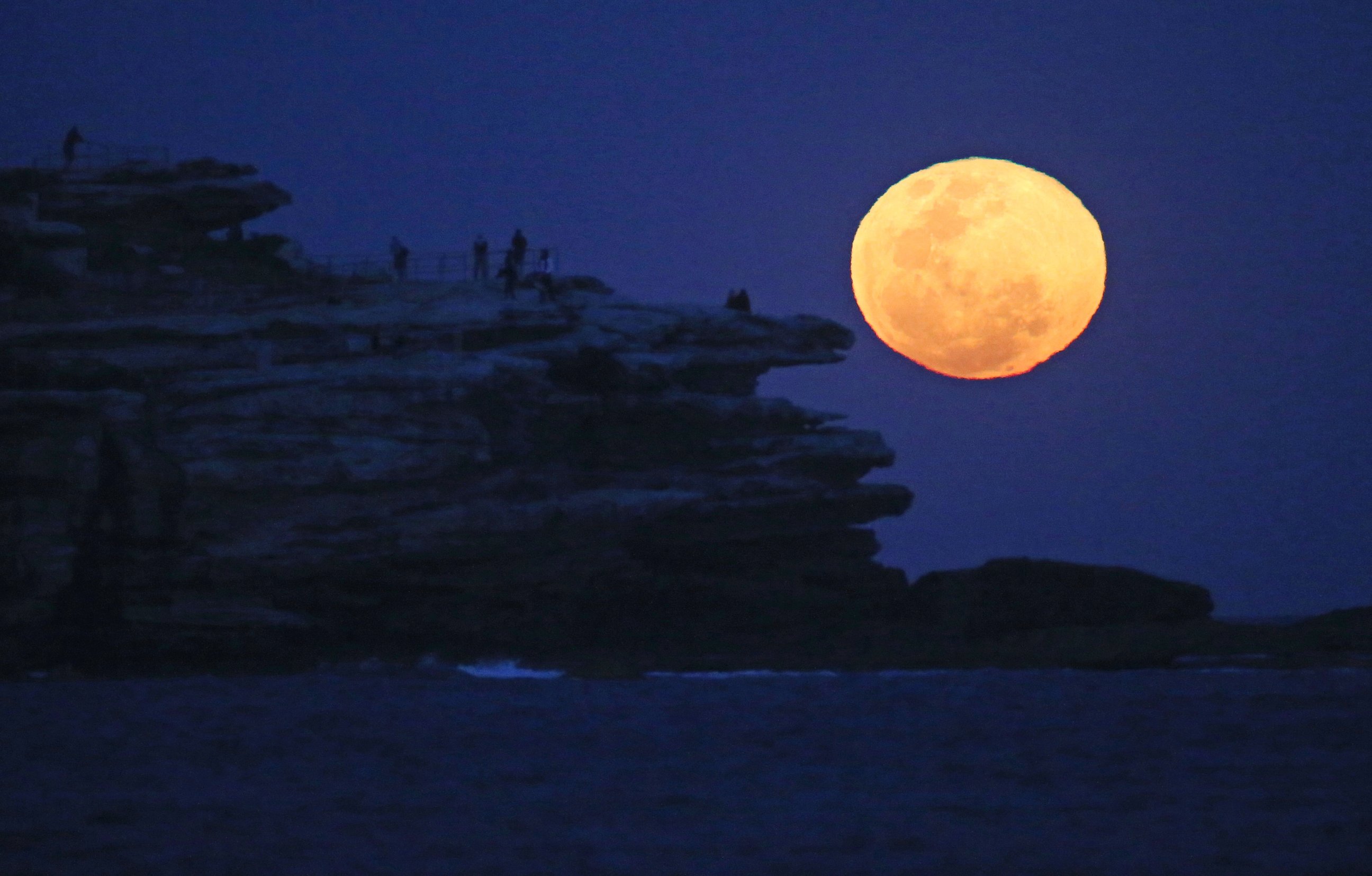 PHOTO: The moon rises with an orange glow as people watch from a rock cliff at Bondi Beach in Sydney, Sept. 9, 2014.