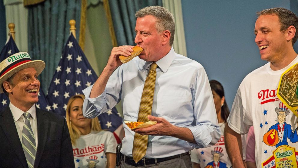 PHOTO: George Shea, left, the official announcer for the Nathan's Hot Dog Eating Contest watches New York City's Mayor Bill De Blasio eating a famous Nathan's hot dog next to Joey "Jaws" Chestnut, right, at City Hall on July 3, 2014 in New York City.