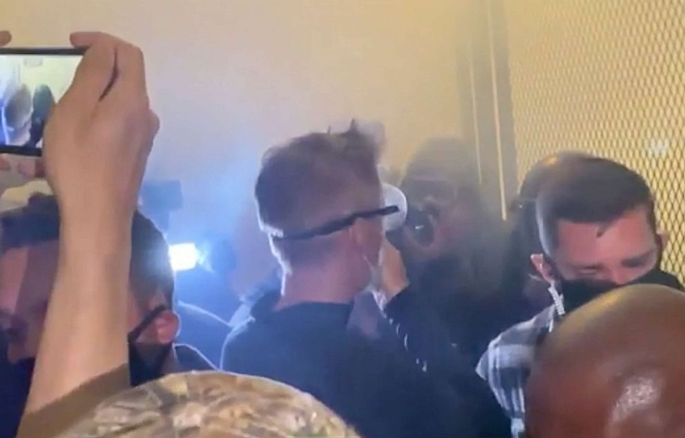 PHOTO: In this image made from video released by Jonathan Maus, Portland Mayor Ted Wheeler, center in black with goggles looking away, stands at a fence guarding a federal courthouse as tear gas drifts by early July 23, 2020, in Portland Oregon.