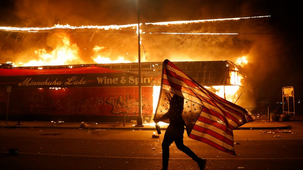 A protester carries a U.S. flag upside down as he walks past a burning building in Minneapolis on May 28, 2020, during a protest over the death of George Floyd,. (AP Photo/Julio Cortez)