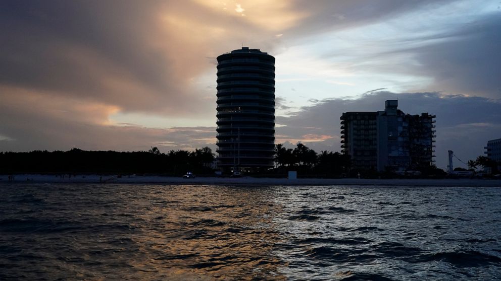 The sun sets over oceanfront buildings, including the partially collapsed Champlain Towers South condo building, right, where demolition experts were preparing to bring down the still-standing portion, Sunday, July 4, 2021, in Surfside, Fla. The deci