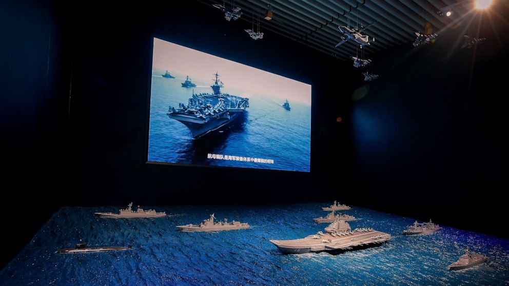 FILE - In this Aug. 1, 2019, file photo, a TV screen showing the U.S. Navy fleet sail in formation near the models of Liaoning aircraft carrier with navy frigates and submarines on display at the military museum in Beijing. China on Wednesday, May 19