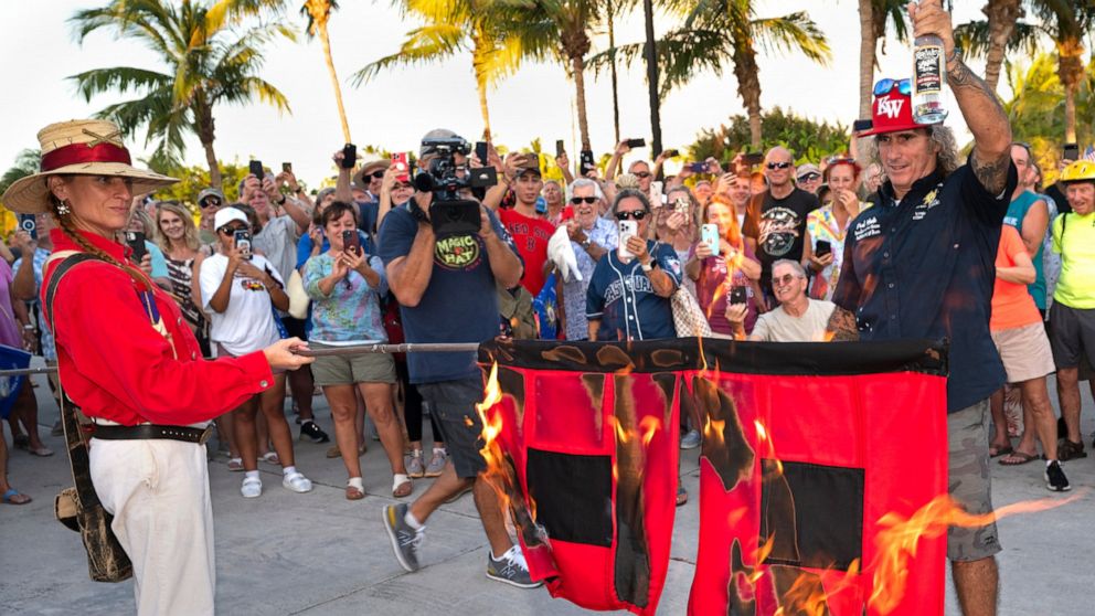 In this photo provided by the Florida Keys News Bureau, Jai Somers, left, holds burning hurricane flags after they were doused with rum and set on fire by Paul Menta, right, to mark the end of the 2022 Atlantic hurricane season Wednesday, Nov. 30, 20