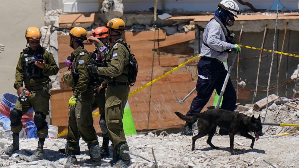 A dog aiding in the search walks past a team of Israeli search and rescue personnel, left, atop the rubble at the Champlain Towers South condo building, where scores of people remain missing one week after it partially collapsed, Friday, July 2, 2021