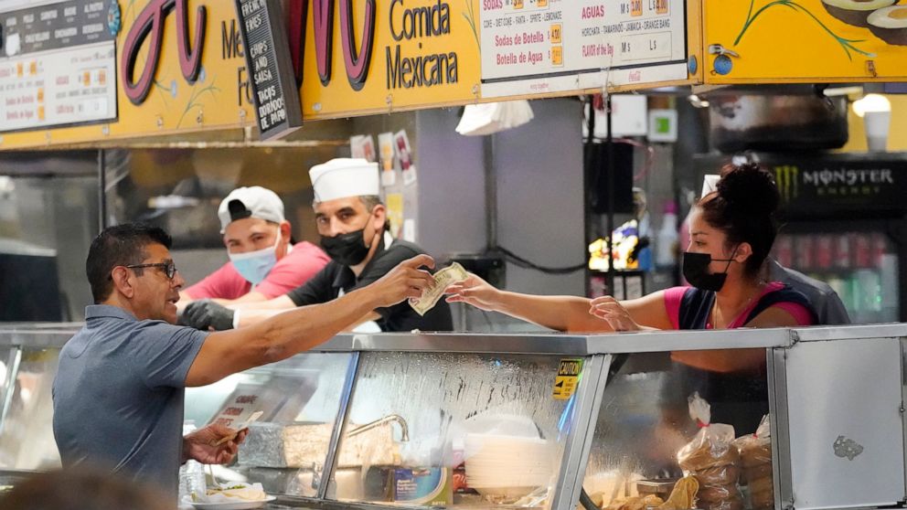 Money is exchanged at a food stand while workers wear face masks inside Grand Central Market on Wednesday, July 13, 2022, in Los Angeles. Falling gas prices gave Americans a slight break from the pain of high inflation last month, though the surge in