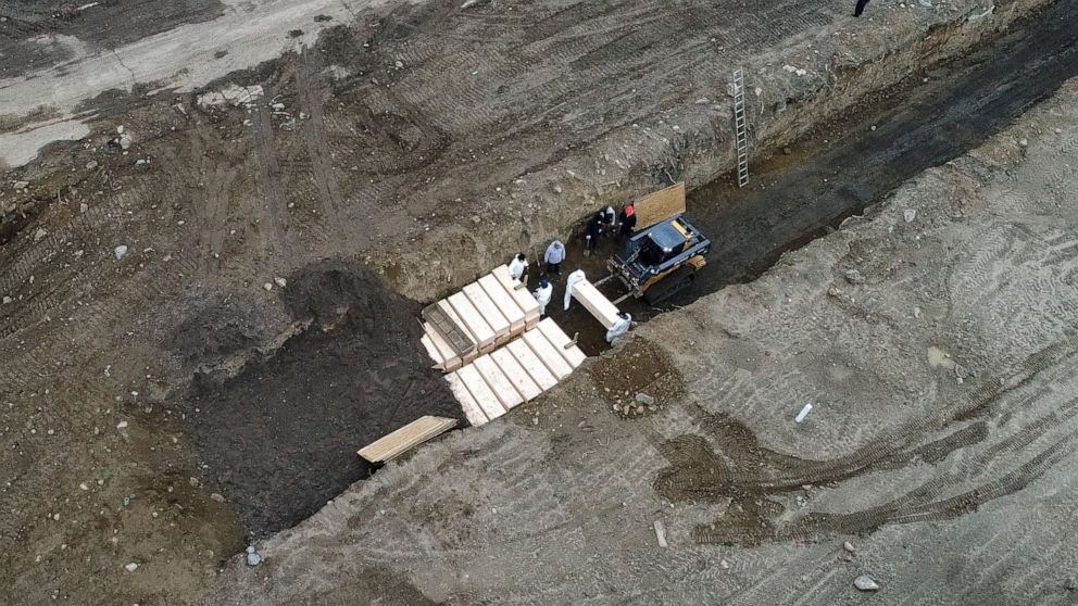 Workers wearing personal protective equipment bury bodies in a trench on Hart Island, Thursday, April 9, 2020, in the Bronx borough of New York. On Thursday, New York City’s medical examiner confirmed that the city has shortened the amount of time it