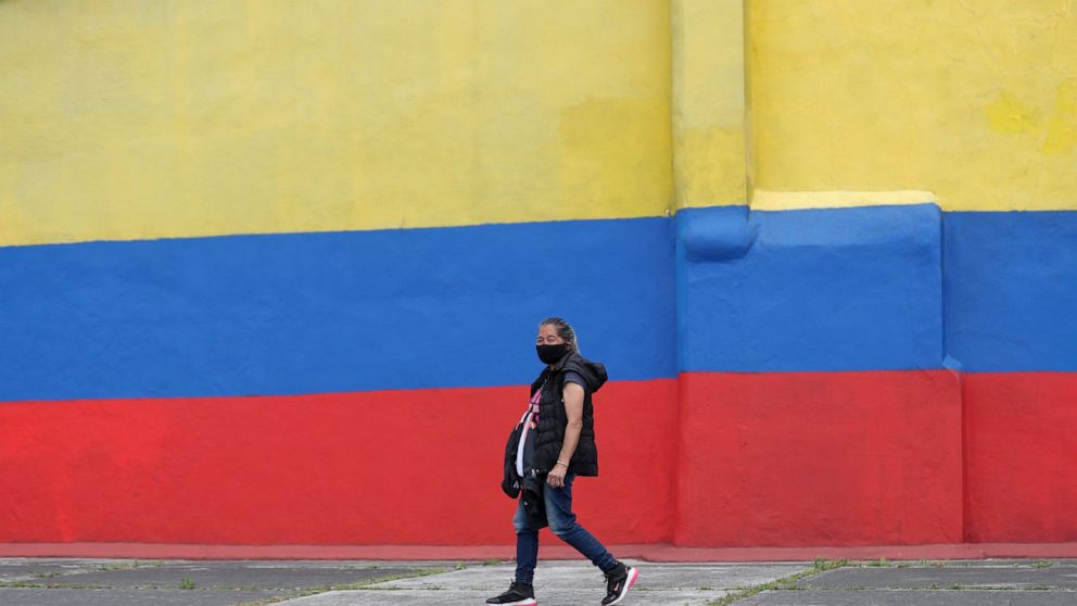 A woman wearing a protective face mask walks next to a house painted with colors of Colombia's flag, during the lockdown ordered by the government in an effort to prevent the spread of the new coronavirus, in Bogota, Colombia, Tuesday, April 14, 2020