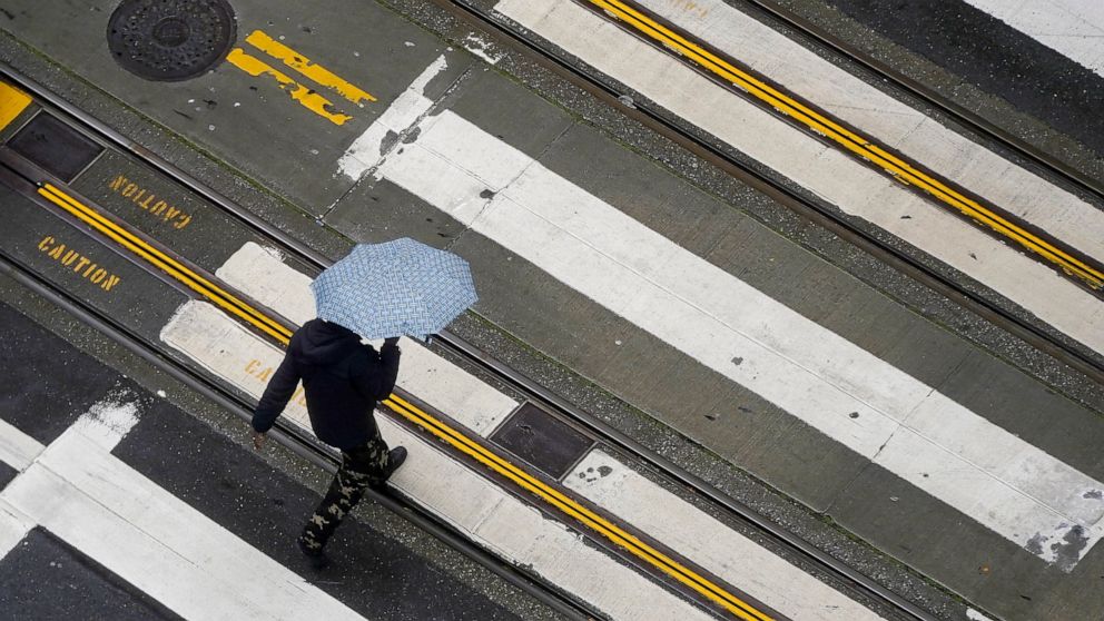 A pedestrian carries an umbrella while crossing a street in San Francisco, Monday, Dec. 13, 2021. A major winter storm hitting Northern California is expected to intensify and bring travel headaches and a threat of localized flooding after an abnorma