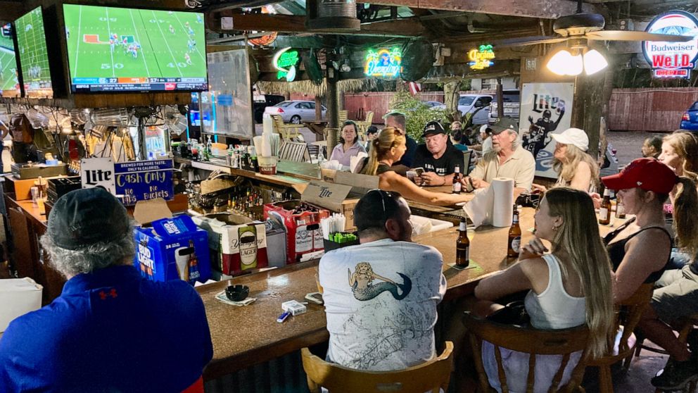 People watch college football games at an outdoor tiki bar under banyan trees in Fort Myers, Fla., on Saturday, Oct. 8, 2022. While some areas are still ravaged by effects of Hurricane Ian, life is returning to normal in other places. (AP Photo/Jay R