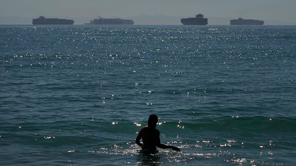 FILE — A man wades through the water at Seal Beach Calif., Friday, Oct. 1, 2021, as container ships waiting to dock at the Ports of Los Angeles and Long Beach are seen in the distance. California state lawmakers held a joint legislative hearing at th