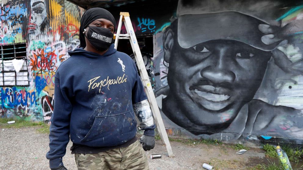 PHOTO: Artist Theo Ponchaveli wears a mask he created that reads, "Runwithmaud", as he poses for a photo in front of a mural that he is painting in the likeness of Ahmaud Arbery in Dallas, Friday, May 8, 2020.