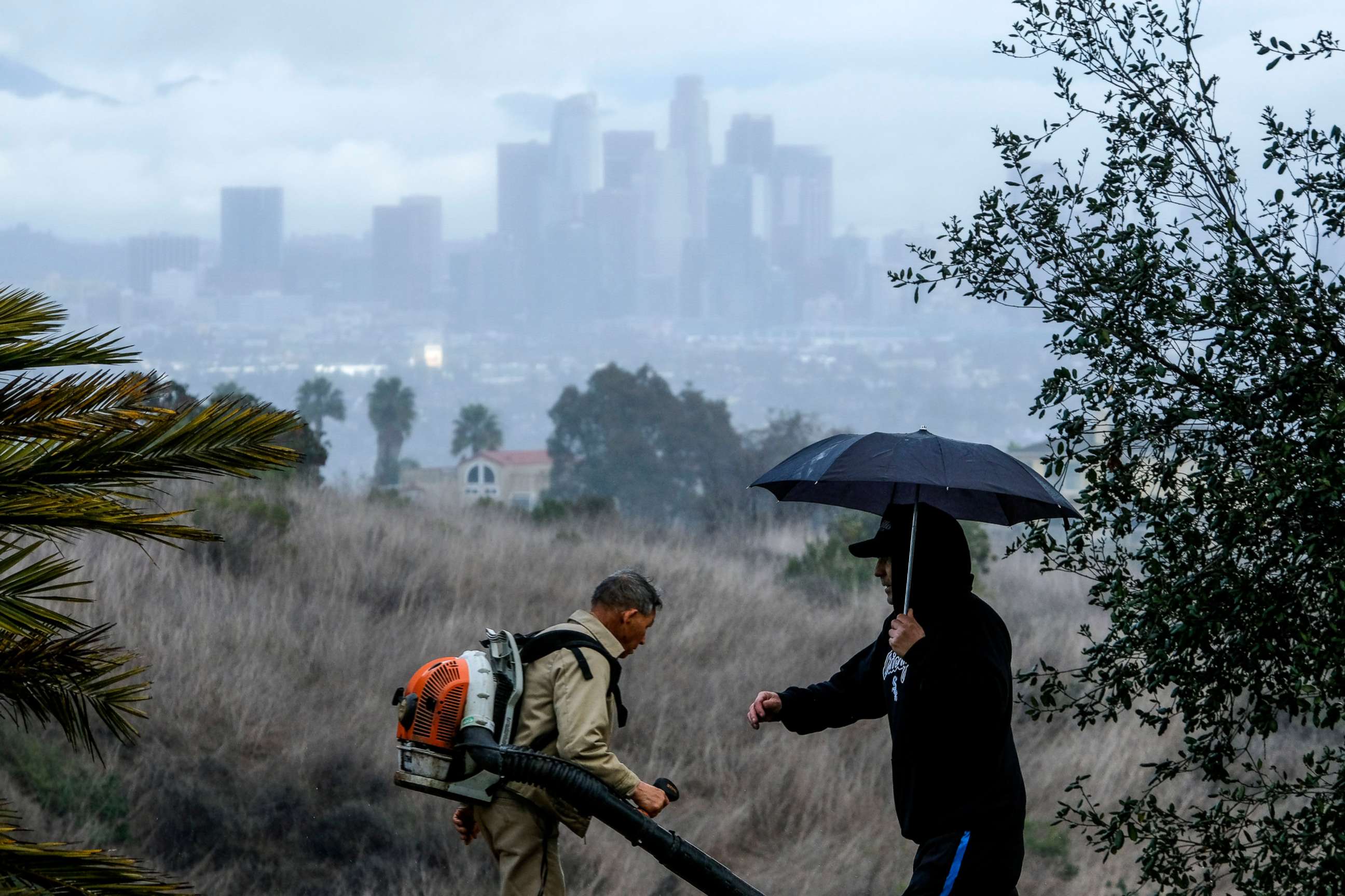 PHOTO: A man with an umbrella walks by a worker in Kenneth Hahn State Recreation Area as the Los Angeles skyline in background, March 10, 2021.