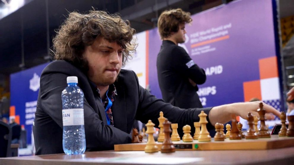 PHOTO: Hans Niemann plays a match at the World Rapid and Blitz Championships as Magnus Carlsen watches other matches from behind.