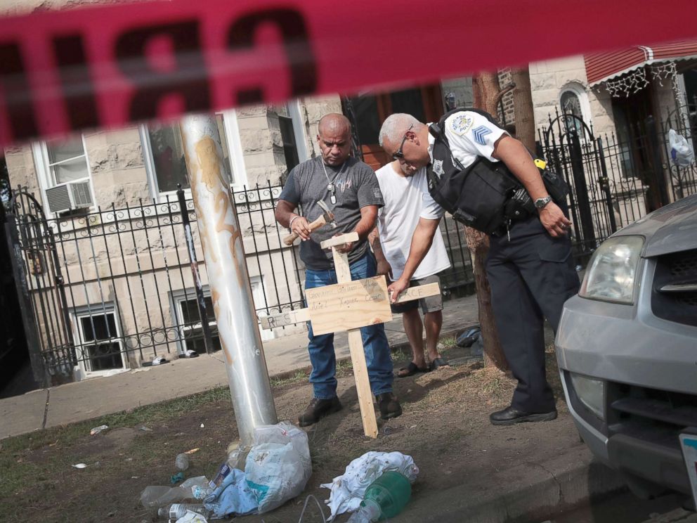 PHOTO: A police officer helps as Omar Acevedo pounds a cross bearing the names of victims into the dirt outside a home where eight people perished, including six children, Aug. 26, 2018, in Chicago.