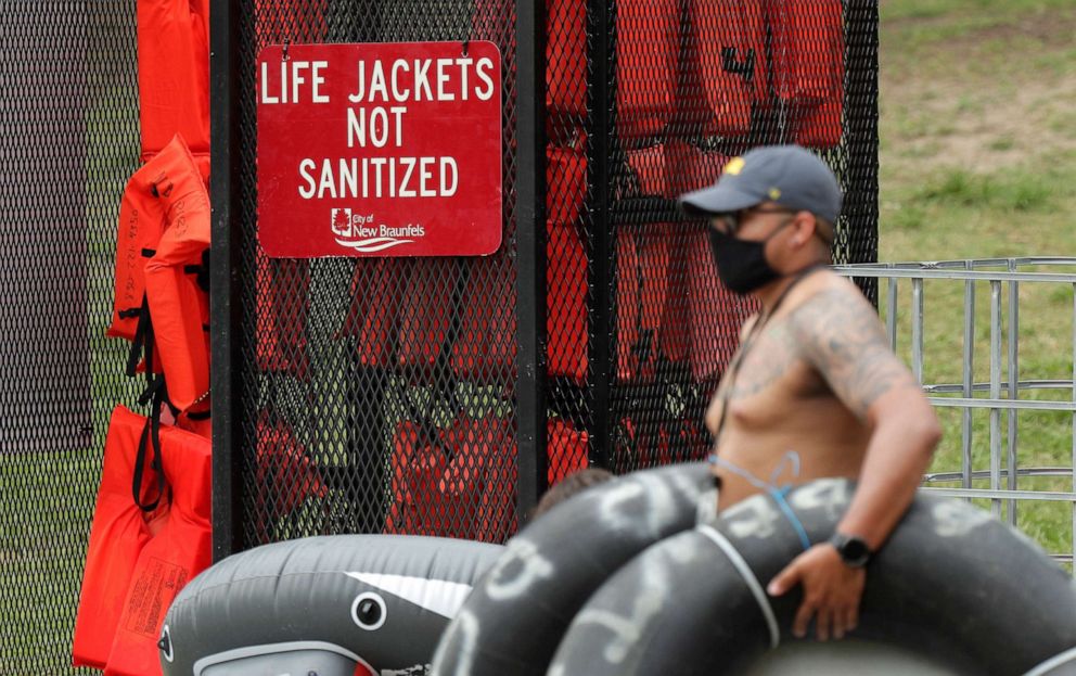 PHOTO: A tuber, wearing a mask to protect against the spread of COVID-19, passes a rack of life jackets as he prepares to float the Comal River, June 25, 2020, in New Braunfels, Texas.