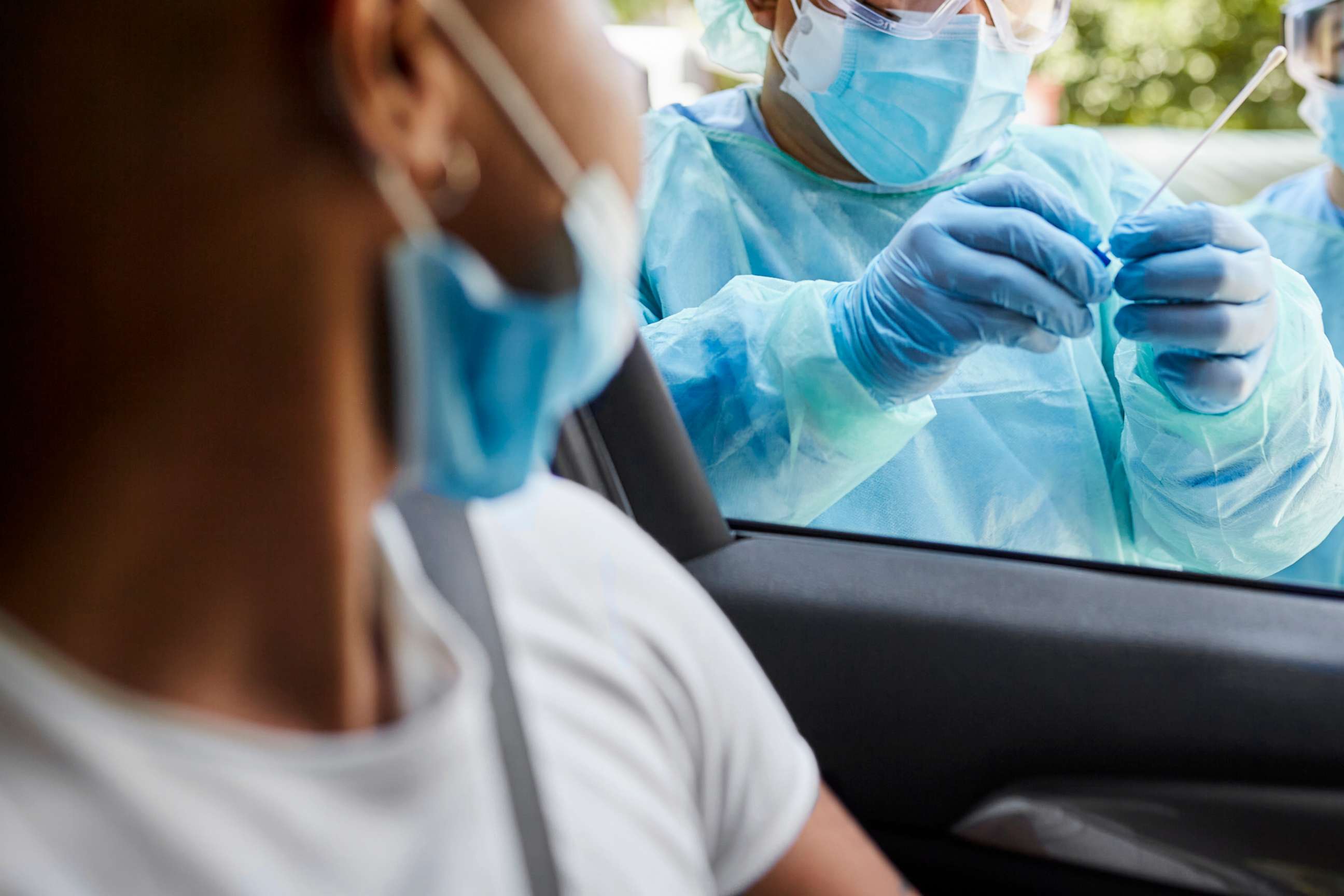 PHOTO: Young man sitting in car watching as hospital team in protective workwear place nasal swab specimen in sterile container.