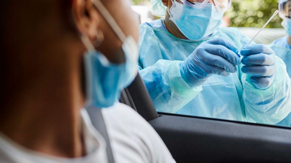 PHOTO: Young man sitting in car watching as hospital team in protective workwear place nasal swab specimen in sterile container.