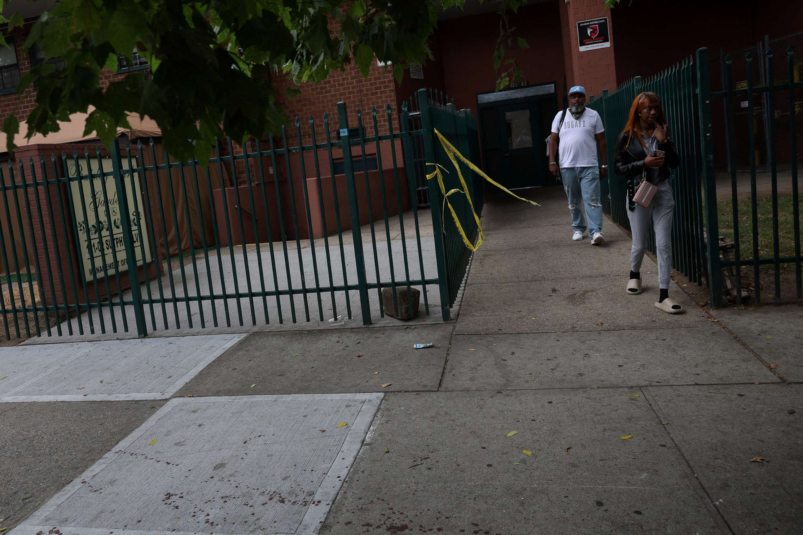 PHOTO: People walk by NYPD police tape and a blood stained sidewalk, where according to police reports a shooting killed one man and injured another Wednesday night outside an apartment building in the South Jamaica section of Queens, July 7, 2022.