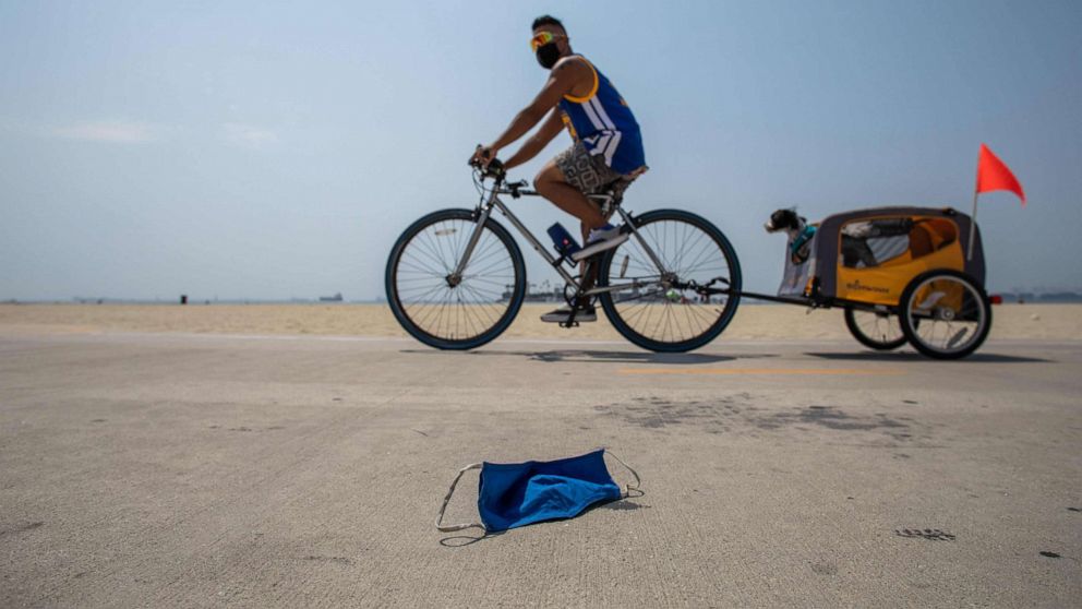 PHOTO: A man wearing a face mask rides his bike towing a dog as a discarded face mask lies along the bike path in Long Beach, California, on Aug. 22, 2020.