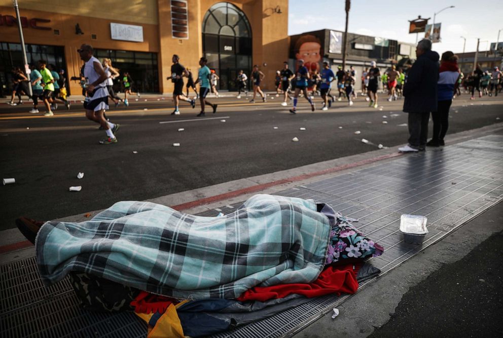 PHOTO: A homeless person sleeps as runners jog past during the Los Angeles Marathon, which was allowed to continue by health officials in spite of coronavirus, on March 8, 2020, in Los Angeles.
