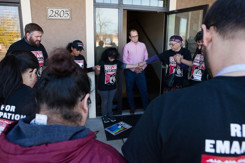 PHOTO: Asamblea members hold a prayer circle outside Representative Jason Lewis's office, while one of his staff members watches.