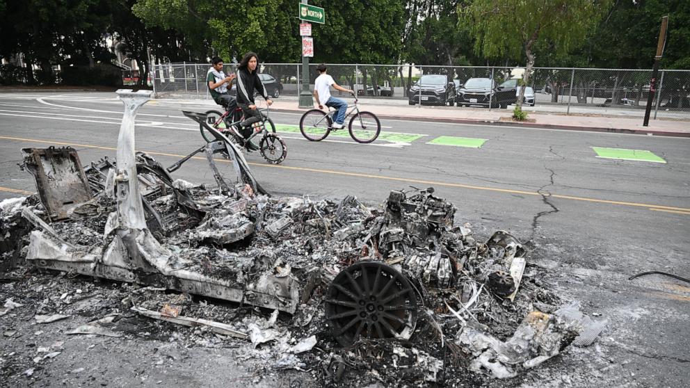 PHOTO: People riding bicycles pass by the charred remains of a burned Waymo vehicle in the street following a night of protests in response to federal immigration operations in Los Angeles, June 9, 2025.