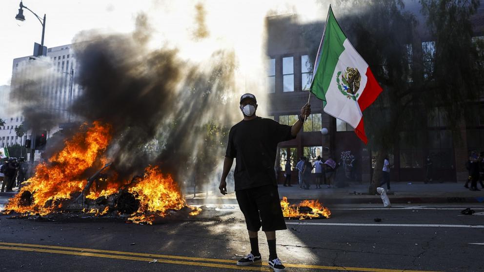 PHOTO: A protester with a Mexican flag looks on as multiple cars burn during immigration raid protests in Los Angeles, June 8, 2025.