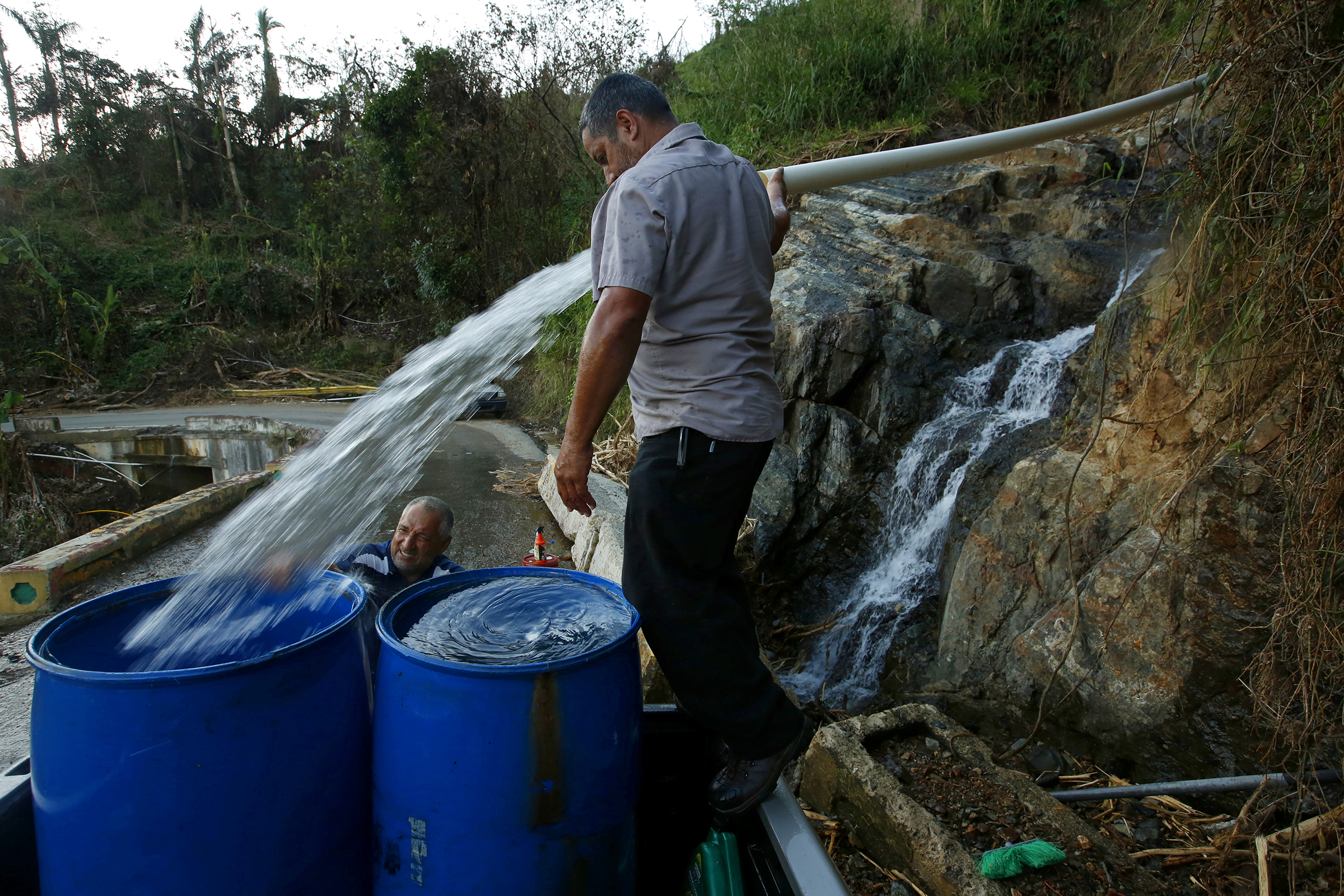 PHOTO: Luis Hernandez, left, gets help from Sergio Rivera, center, with filling drums with spring water for washing in Jayuya, Puerto Rico, the public water system isn't working full-time, so many collect water from the mountain springs, Oct. 4, 2017.