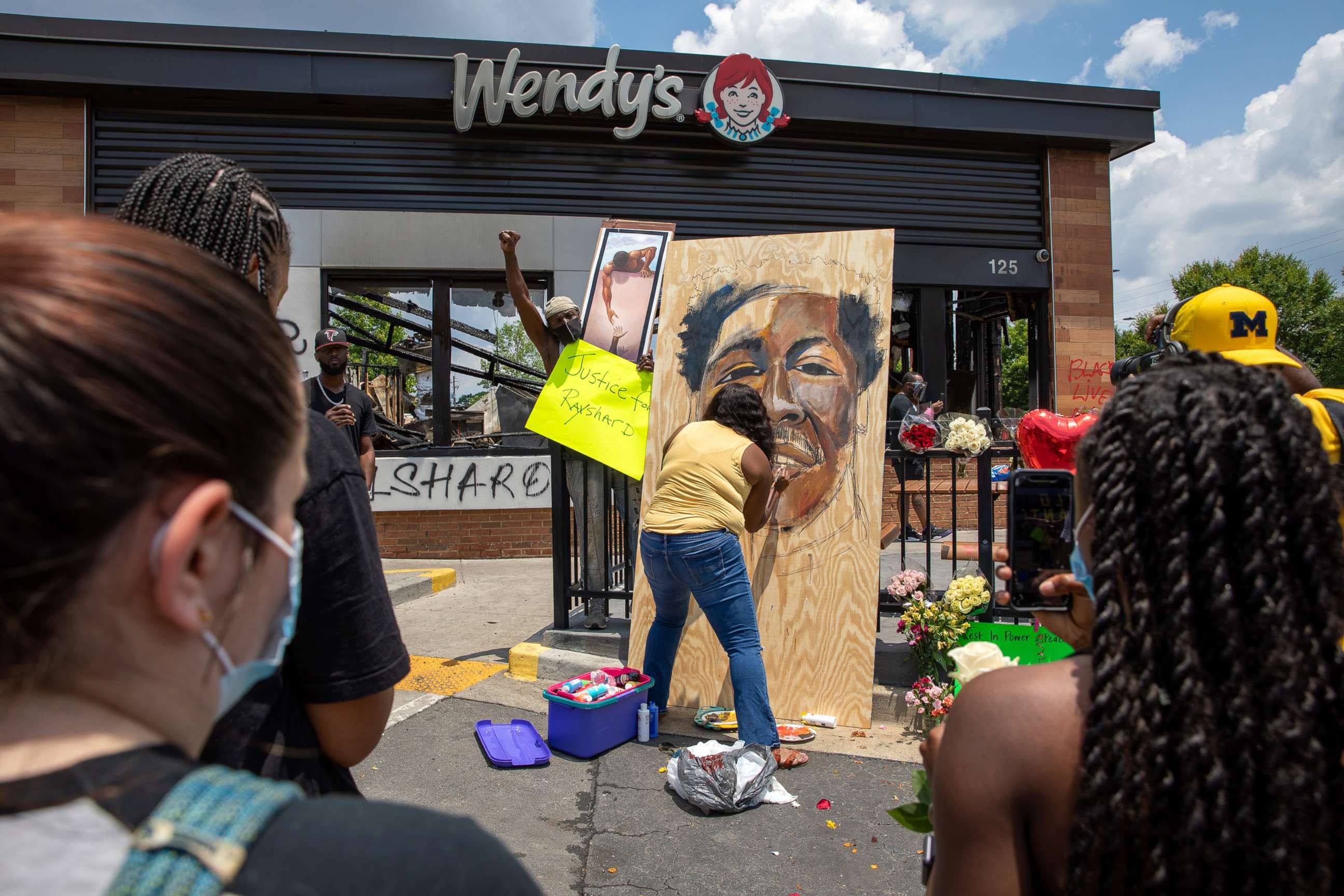 PHOTO: People gather at a memorial as a woman paints a portrait of Rayshard Brooks at the site of a Wendy's restaurant set ablaze overnight on June 14, 2020 in Atlanta. 