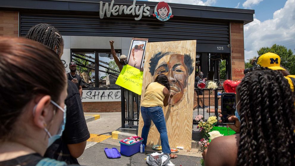 PHOTO: People gather at a memorial as a woman paints a portrait of Rayshard Brooks at the site of a Wendy's restaurant set ablaze overnight on June 14, 2020 in Atlanta. 