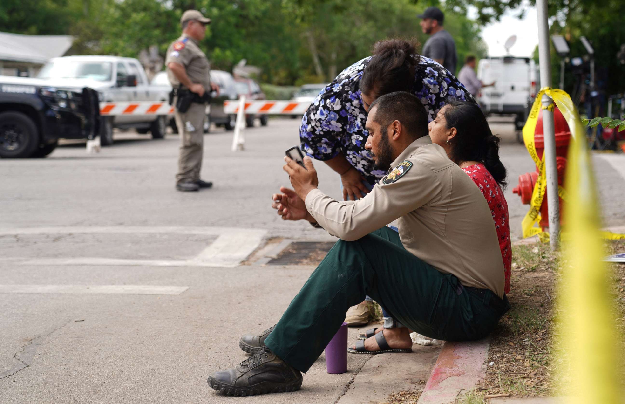 PHOTO: A sheriff checks his phone as he sits on the sidewalk with two women outside Robb Elementary School as state troopers monitor the area in Uvalde, Texas, May 24, 2022.