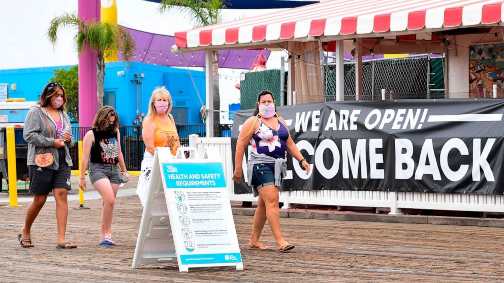 PHOTO: People wearing face masks walk past a health and safety guidelines board and an open restaurant on the pier in Santa Monica, California, on June 26, 2020.