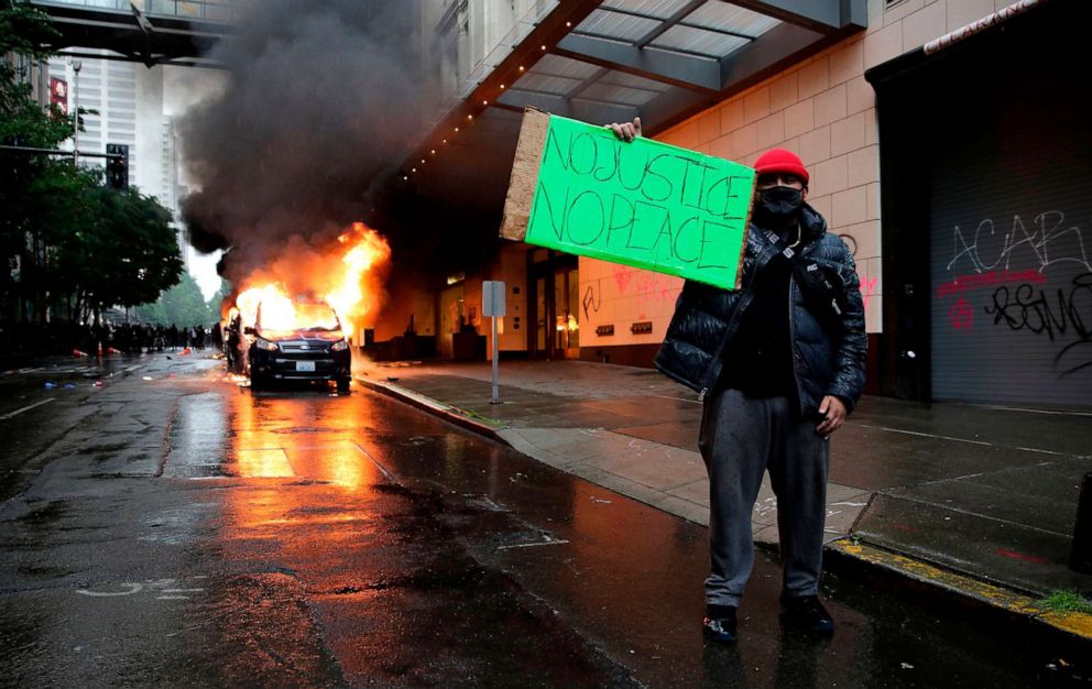 PHOTO: A man holds a sign in front of burning vehicles following demonstrations protesting the death of George Floyd, in Seattle, May 30, 2020.