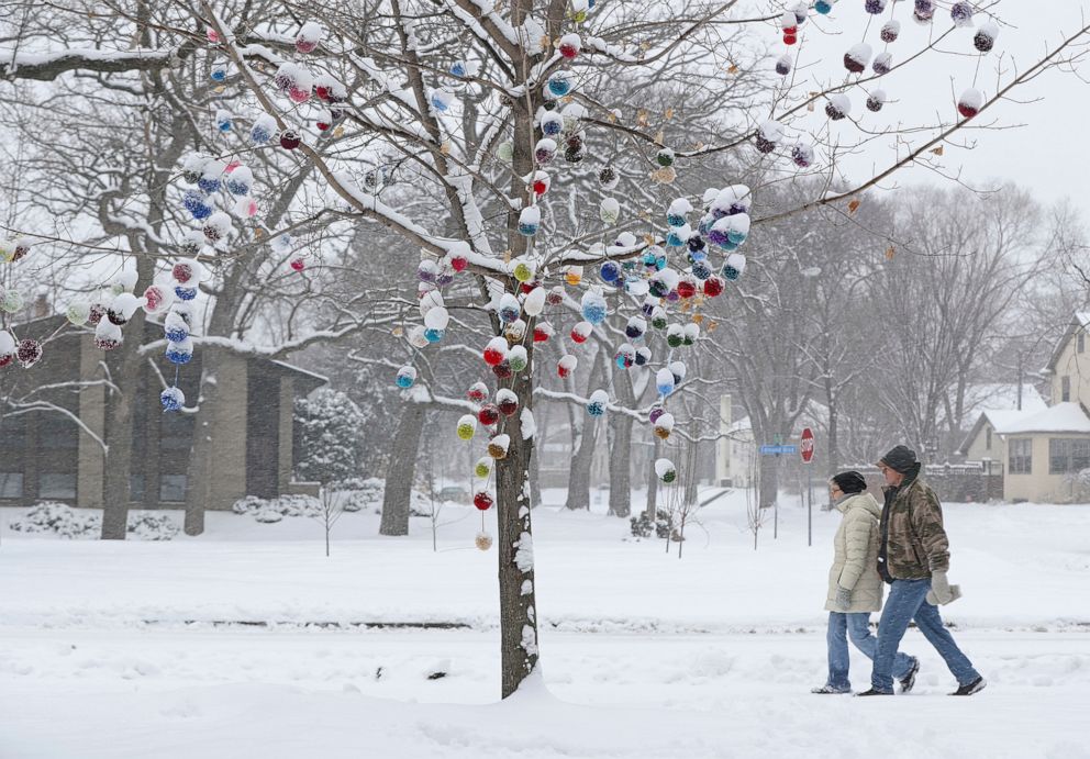 PHOTO: Snow covers the tops of knitted yarn pom-poms decorating a tree along West River Road as a couple walks by in Minneapolis, April 3, 2021.