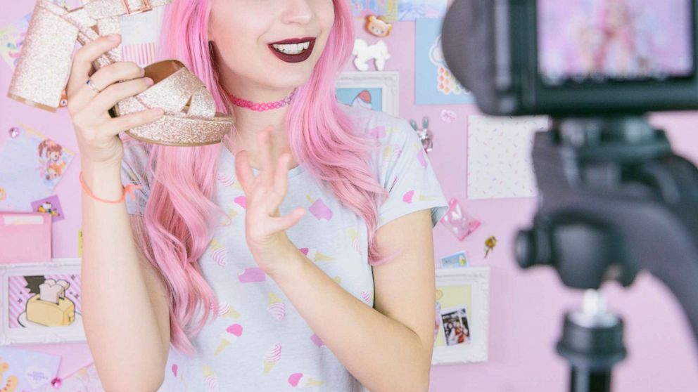 PHOTO: A staged photo of someone portraying a Social media influencer reviewing footwear, she is vlogging about women's fashion and filming herself at home on a video camera.