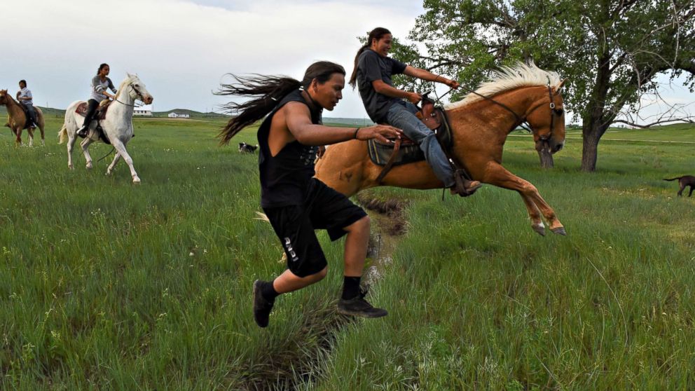 Mahto In The Woods jumps a small creek on foot while his cousin Jayden Lookinghorse jumps over on his horse on the Cheyenne River Reservation in Green Grass, South Dakota, May 31, 2018.