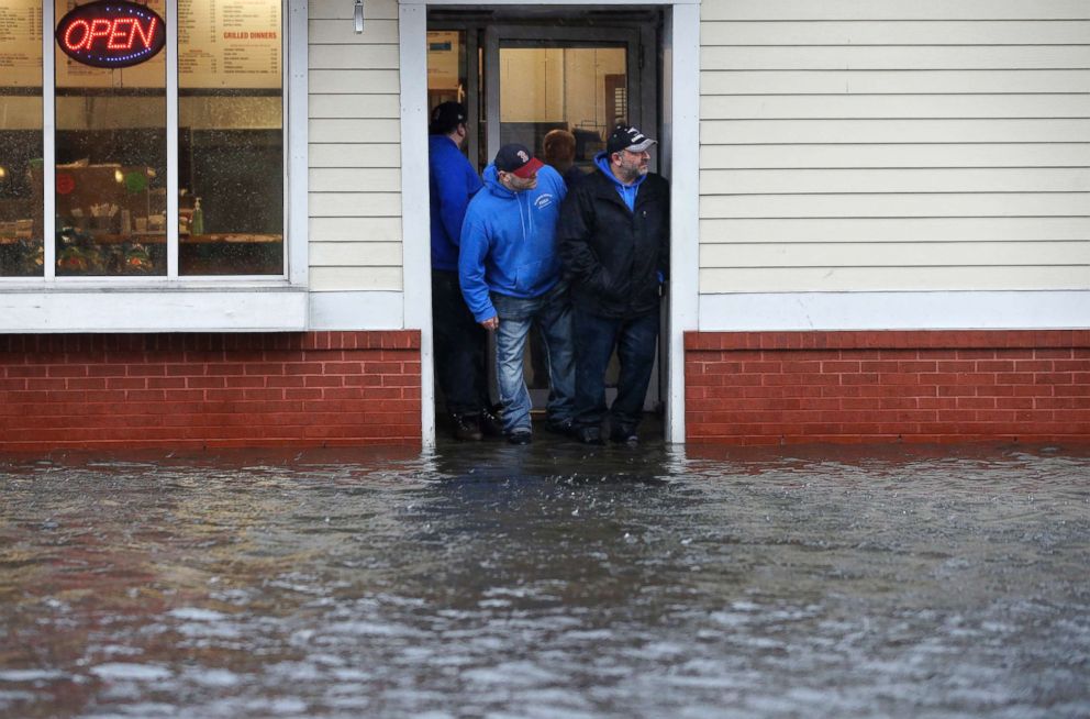 PHOTO: People stand at the entrance to a pizza shop as water floods a street, in Scituate, Mass., March 2, 2018.