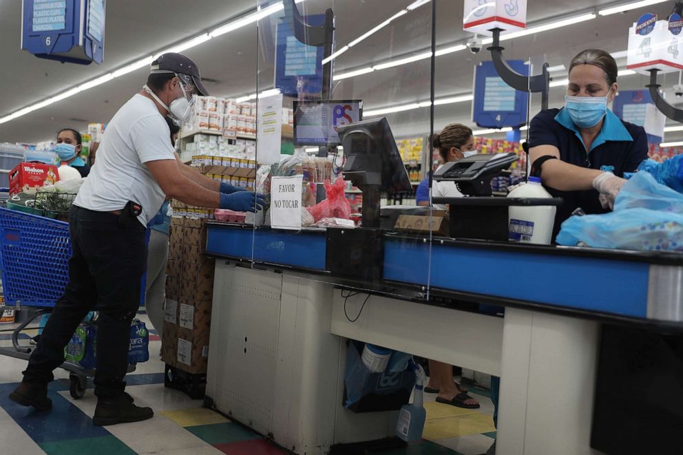PHOTO: Lay Guzman stands behind a partial protective plastic screen and wears a mask and gloves as she works as a cashier at the Presidente Supermarket on April 13, 2020 in Miami, Florida.