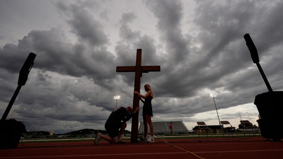 PHOTO: Dan Beazley, of Michigan, left, reacts as he holds a large cross with Abigail Smithson during a vigil for flooding victims at Tivy Antler Stadium, July 9, 2025, in Kerrville, Texas.