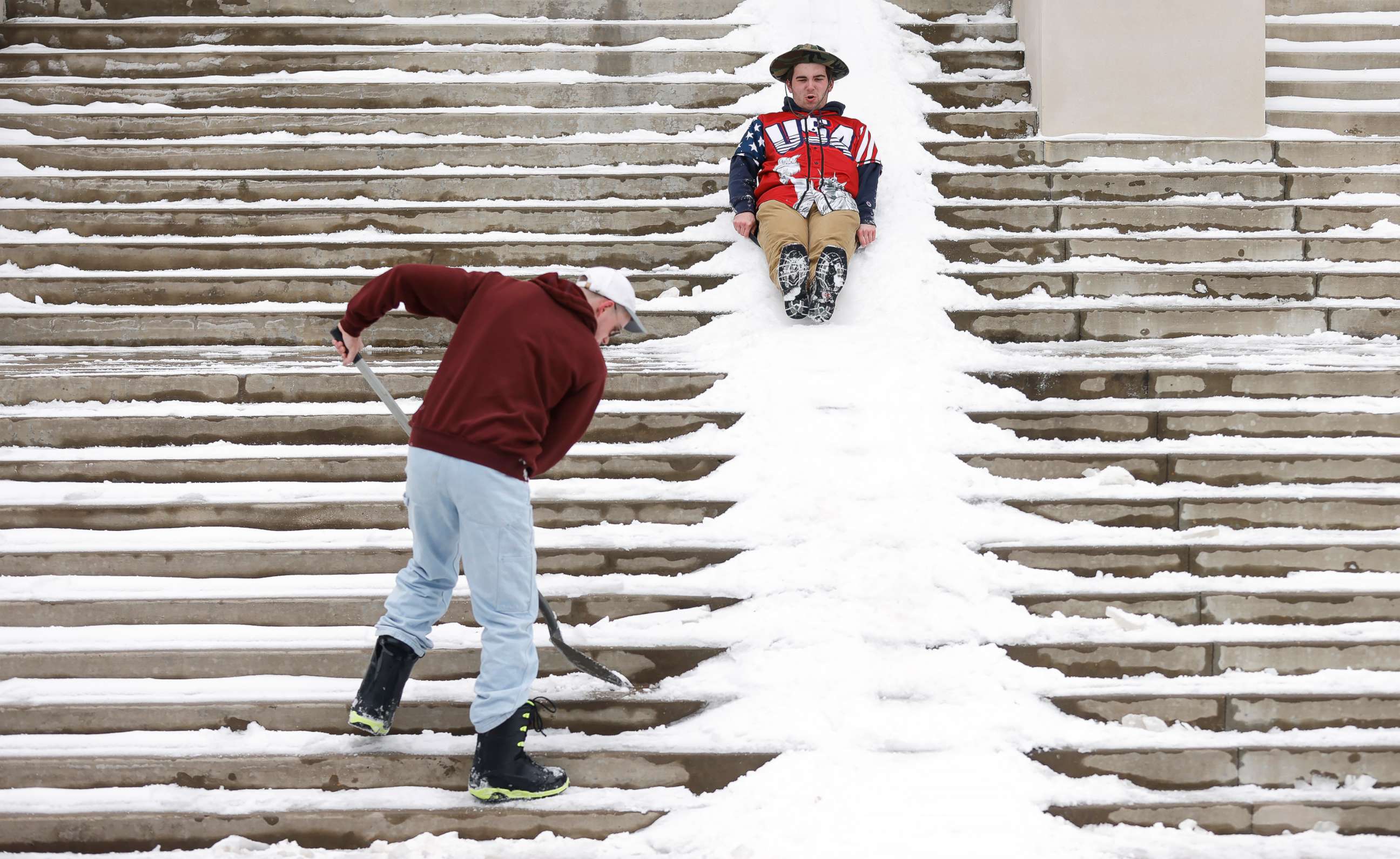 PHOTO: Jimmy Pavelich sleds down the steps of the Mary Couts Burnett Library as Michael Manson shovels snow at Texas Christian University after a snow storm on Feb. 17, 2021, in Fort Worth, Texas.