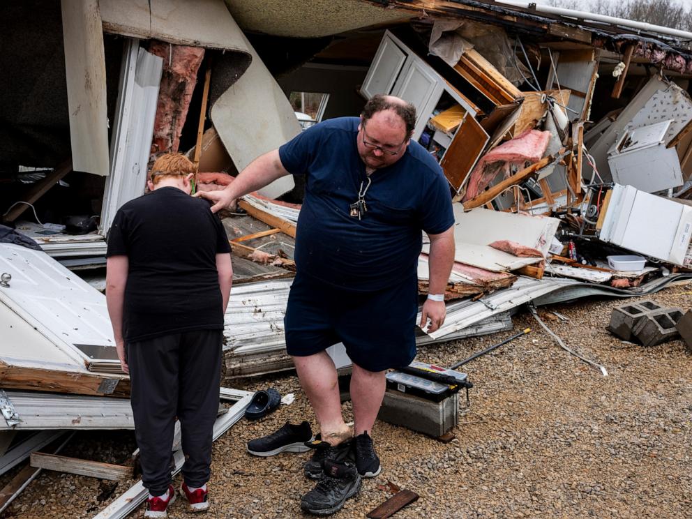 PHOTO: Justin Stievenart, right, uses his son, Jayson, 11, for balance while switching shoes outside of his destroyed home in Harmony Hills trailer park on March 15, 2025, in Poplar Bluff, Missouri.