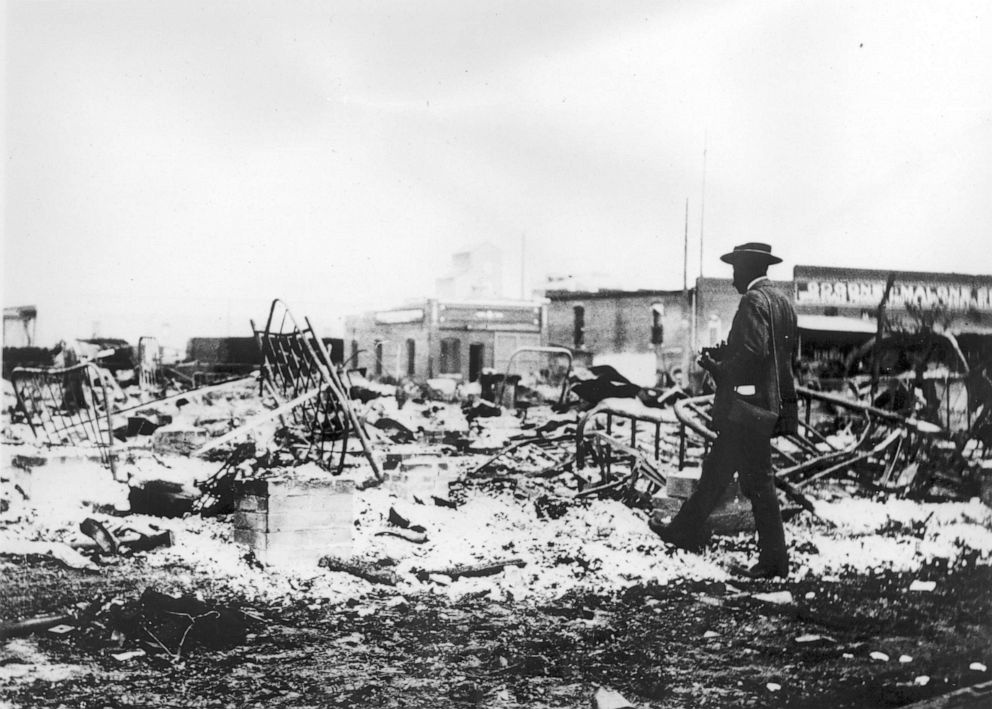 PHOTO: Photograph of an African-American man with a camera looking at the skeletons of iron beds which rise above the ashes of a burned-out block after the Tulsa Race Massacre, Tulsa, Okla., 1921.