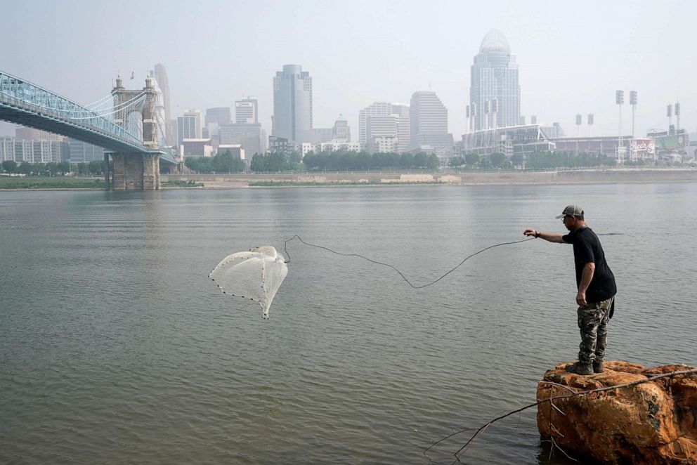 PHOTO: A man tosses a fishing net into the Ohio River as smoke from wildfires is visible over downtown Cincinnati, June 28, 2023.